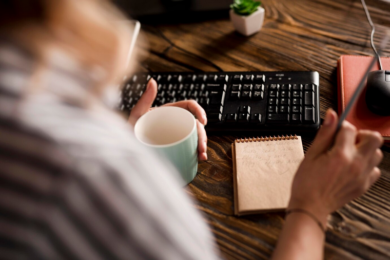 Человек держит светло-зеленую кружку и работает за столом с клавиатурой и блокнотом.