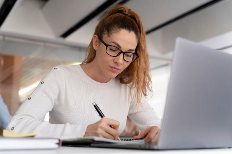 Женщина в очках пишет в блокноте, работая на ноутбуке.